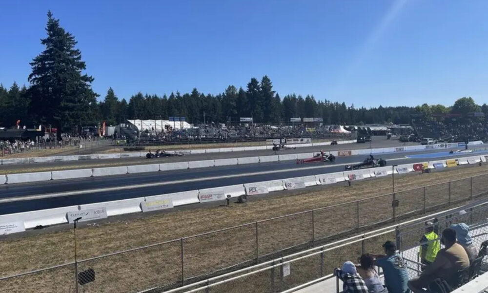 Drag strip with multiple race cars on the track as spectators sit in the bleachers under a clear blue sky.