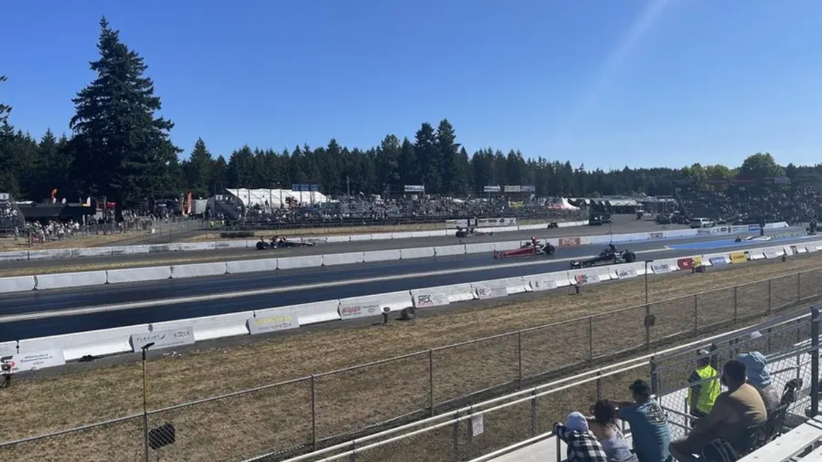 Drag strip with multiple race cars on the track as spectators sit in the bleachers under a clear blue sky.