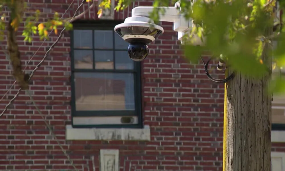 Outdoor dome security camera mounted on a white bracket beside a brick building, monitoring the area outside a window.