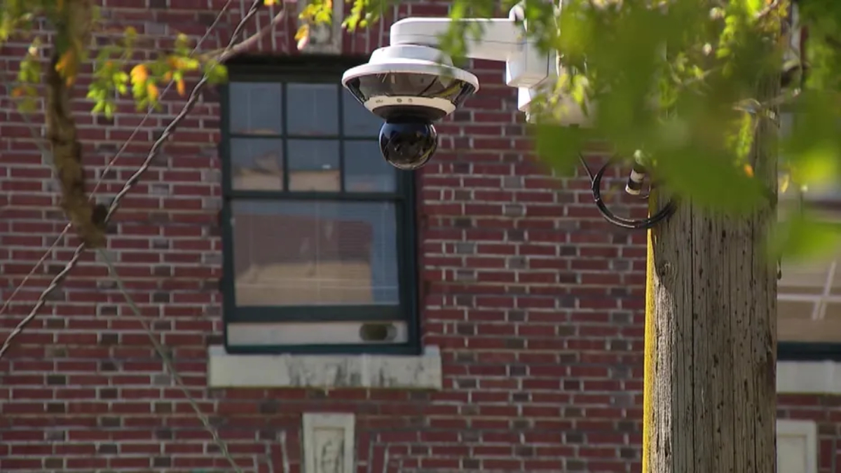 Outdoor dome security camera mounted on a white bracket beside a brick building, monitoring the area outside a window.