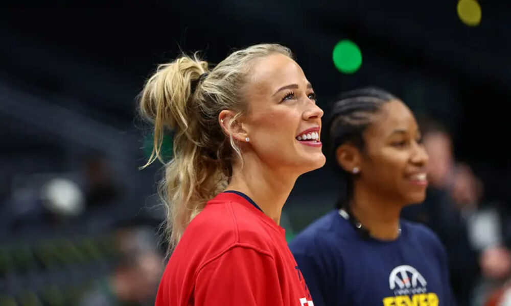 Two smiling women in a sports setting: foreground is a blonde woman in a red shirt with a ponytail, background shows a teammate in a navy shirt.