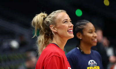 Two smiling women in a sports setting: foreground is a blonde woman in a red shirt with a ponytail, background shows a teammate in a navy shirt.