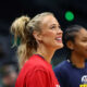 Two smiling women in a sports setting: foreground is a blonde woman in a red shirt with a ponytail, background shows a teammate in a navy shirt.