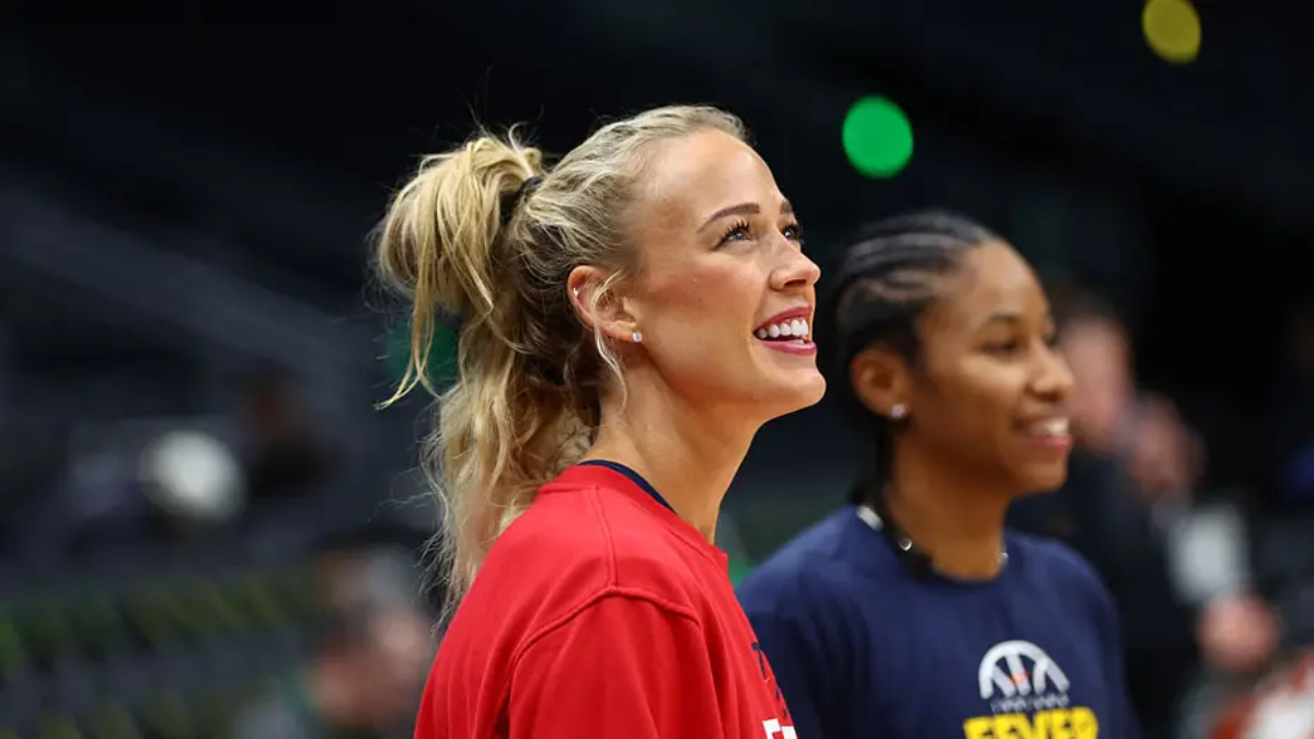 Two smiling women in a sports setting: foreground is a blonde woman in a red shirt with a ponytail, background shows a teammate in a navy shirt.