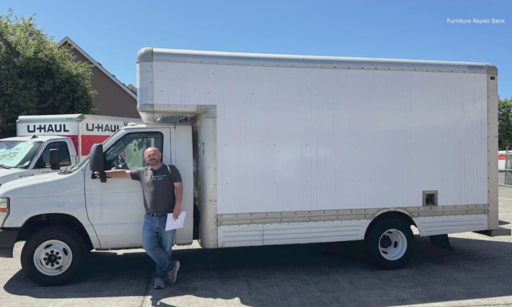 Man leaning on a large white box truck in a parking lot, with U-Haul vans in the background on a sunny day.
