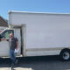 Man leaning on a large white box truck in a parking lot, with U-Haul vans in the background on a sunny day.