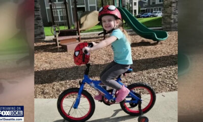 Young girl wearing a Spider-Man helmet rides a blue kid's bicycle with training wheels at a playground, smiling.