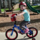 Young girl wearing a Spider-Man helmet rides a blue kid's bicycle with training wheels at a playground, smiling.