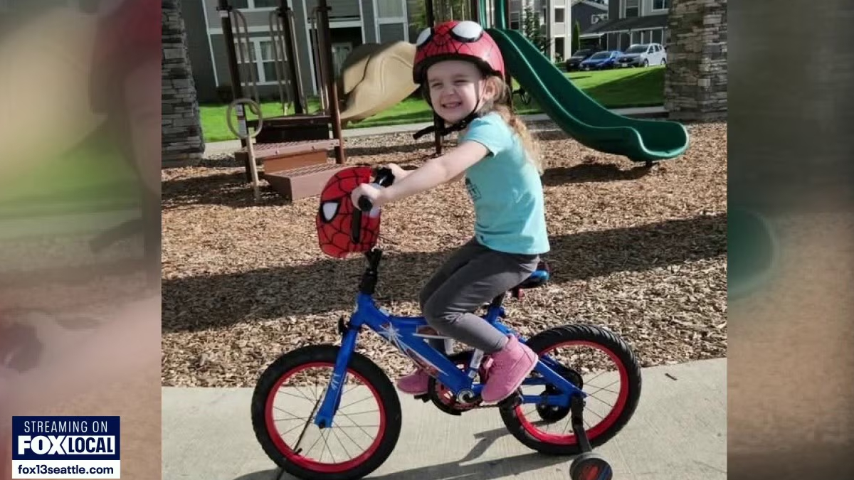 Young girl wearing a Spider-Man helmet rides a blue kid's bicycle with training wheels at a playground, smiling.