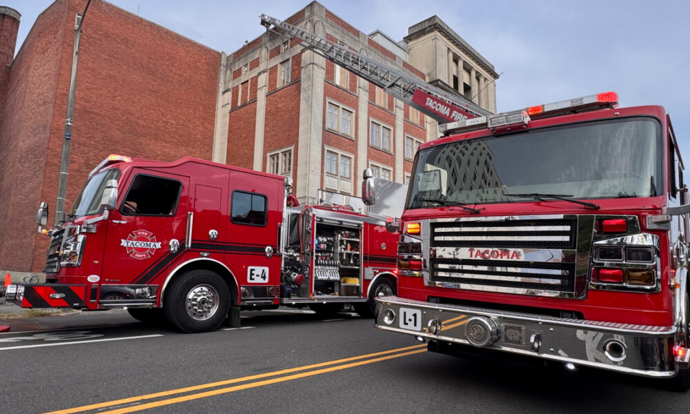 Two red fire engines are parked on a city street with a ladder truck extending its ladder toward a brick building, ready for operation.