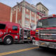 Two red fire engines are parked on a city street with a ladder truck extending its ladder toward a brick building, ready for operation.