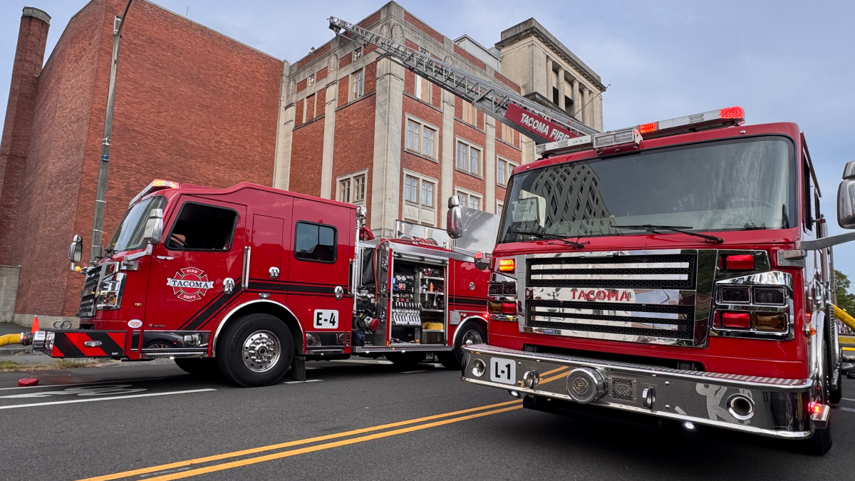 Two red fire engines are parked on a city street with a ladder truck extending its ladder toward a brick building, ready for operation.