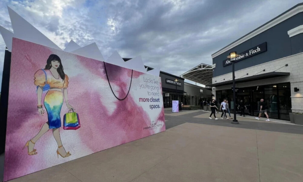 Large pink mural at a shopping center showing an illustrated woman with a colorful bag, outside a store complex including Abercrombie & Fitch.