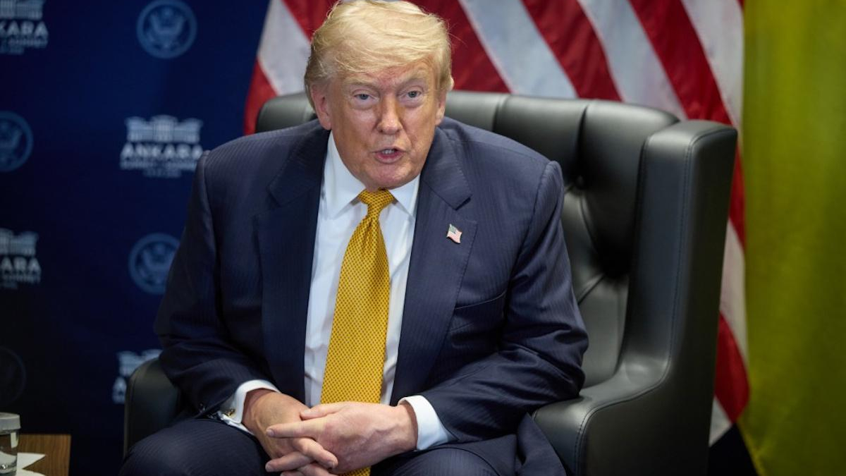 Man in a navy suit and yellow tie sits in a leather chair, speaking, with an American flag and blue backdrop in the background.