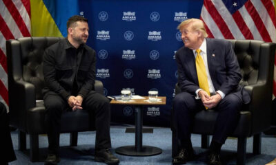 Two men sit in black leather chairs facing each other in a formal meeting, with a blue backdrop featuring Ankara logos and flags on either side.