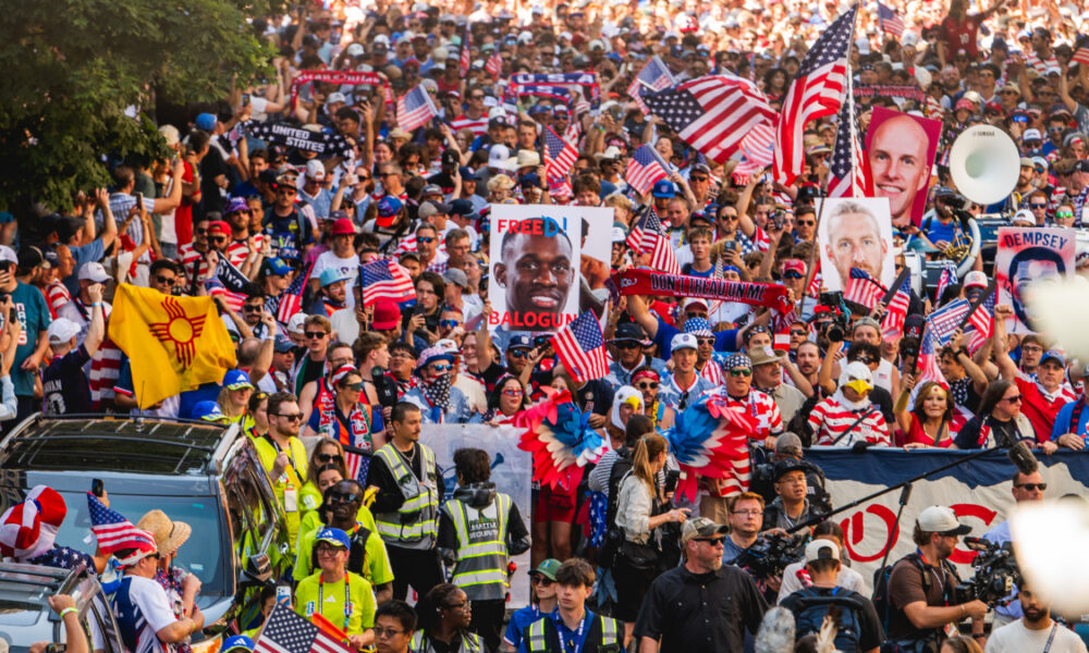 Dense crowd at a patriotic protest, waving American flags and holding signs like 'Free Balogun' amid security personnel at the front.