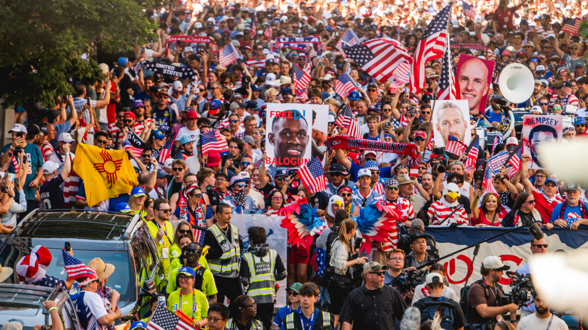 Dense crowd at a patriotic protest, waving American flags and holding signs like 'Free Balogun' amid security personnel at the front.