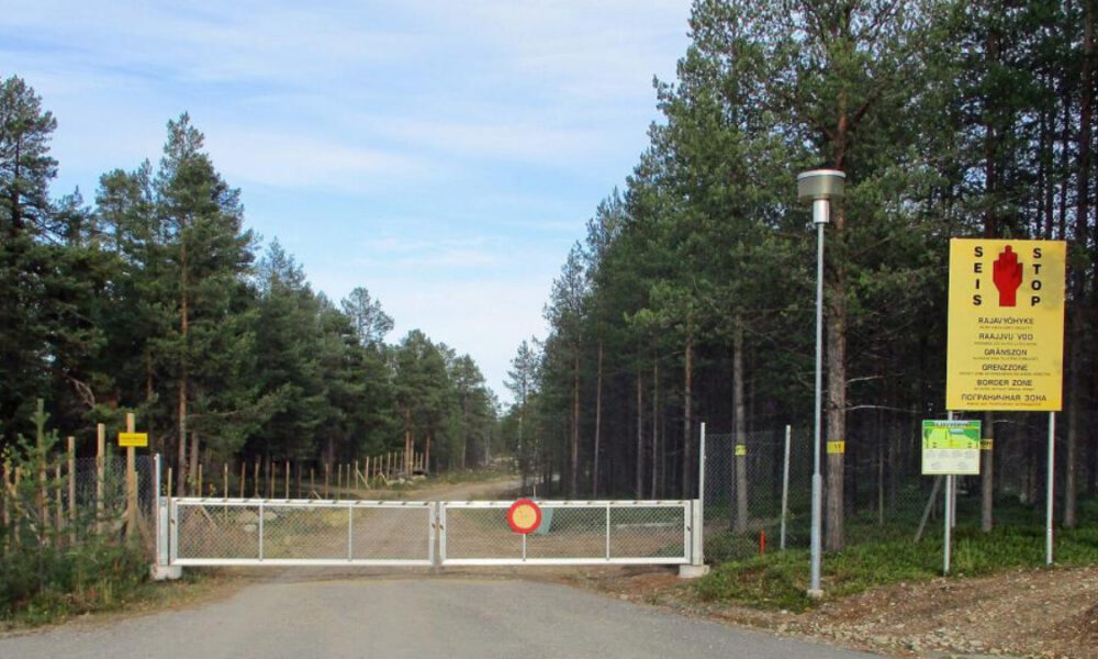 Gate blocks a forest dirt road with a red stop sign on the gate and a tall yellow multilingual warning sign to the right, indicating no entry.