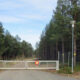 Gate blocks a forest dirt road with a red stop sign on the gate and a tall yellow multilingual warning sign to the right, indicating no entry.