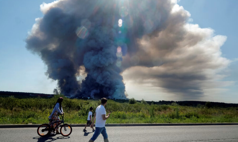 Distant large smoke plume rising from a field, while a cyclist and two pedestrians pass along a sunny roadside in the foreground.