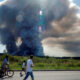 Distant large smoke plume rising from a field, while a cyclist and two pedestrians pass along a sunny roadside in the foreground.