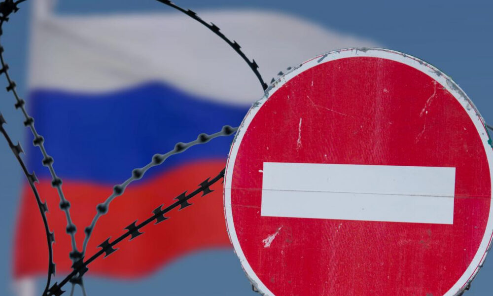 Red no-entry sign in front of barbed wire with the blurred Russian flag in the background, indicating border restriction.