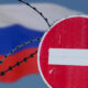 Red no-entry sign in front of barbed wire with the blurred Russian flag in the background, indicating border restriction.