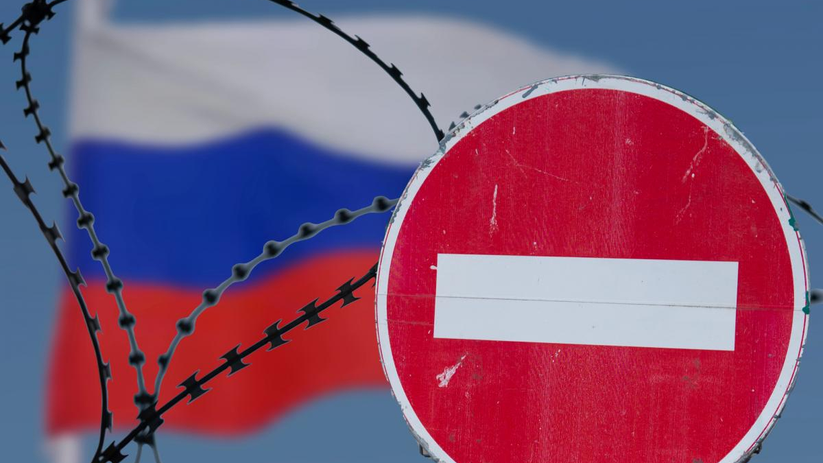 Red no-entry sign in front of barbed wire with the blurred Russian flag in the background, indicating border restriction.