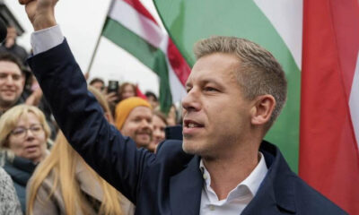 Man in a dark blazer raises his fist at a political rally, surrounded by supporters and flags in the background.