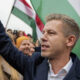 Man in a dark blazer raises his fist at a political rally, surrounded by supporters and flags in the background.
