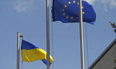 European Union flag with a circle of yellow stars waving next to the Ukrainian flag on flagpoles against a clear blue sky, outdoors.