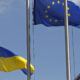 European Union flag with a circle of yellow stars waving next to the Ukrainian flag on flagpoles against a clear blue sky, outdoors.