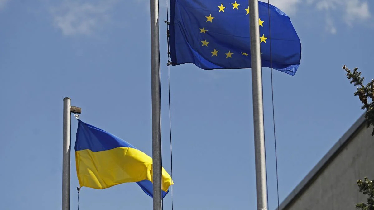 European Union flag with a circle of yellow stars waving next to the Ukrainian flag on flagpoles against a clear blue sky, outdoors.