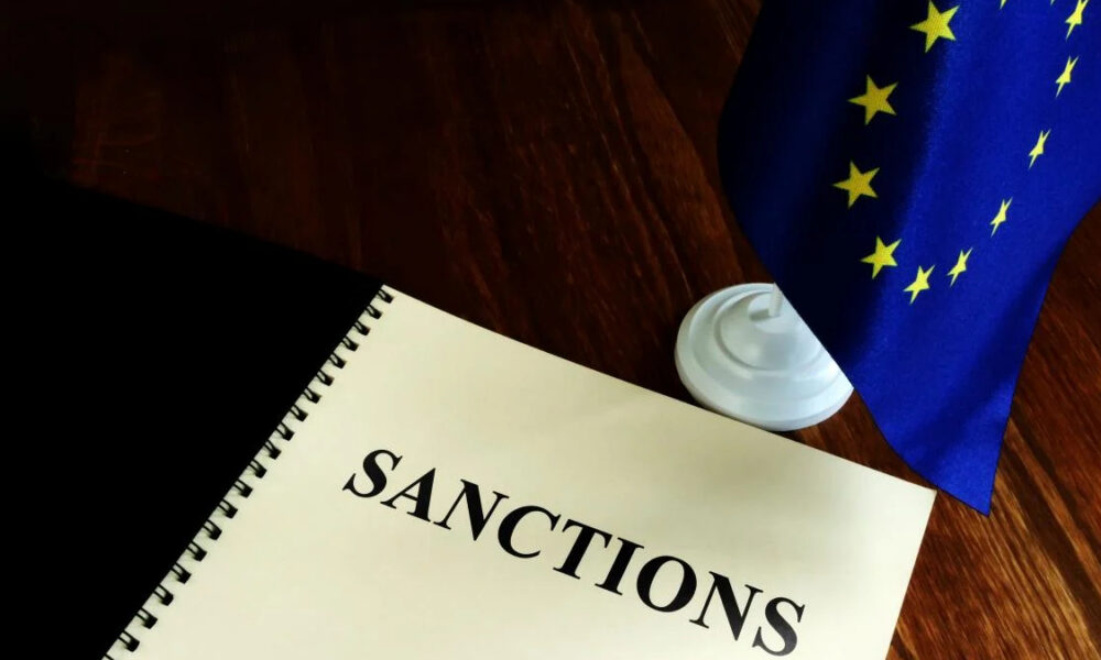 Notebook opened to the word 'SANCTIONS' beside a small EU flag on a wooden table, signaling diplomacy or sanctions policy.