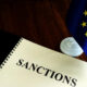 Notebook opened to the word 'SANCTIONS' beside a small EU flag on a wooden table, signaling diplomacy or sanctions policy.