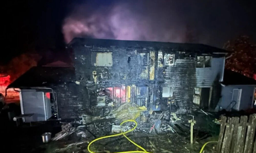 Two-story home severely burned at night with charred exterior, smoke rising, and scattered debris in the yard, including a yellow hose in the foreground