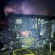 Two-story home severely burned at night with charred exterior, smoke rising, and scattered debris in the yard, including a yellow hose in the foreground