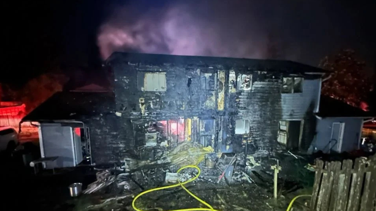 Two-story home severely burned at night with charred exterior, smoke rising, and scattered debris in the yard, including a yellow hose in the foreground