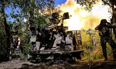 Soldiers in camouflage guard an armored vehicle as its cannon fires in a forest clearing, flames and smoke behind them.