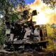 Soldiers in camouflage guard an armored vehicle as its cannon fires in a forest clearing, flames and smoke behind them.