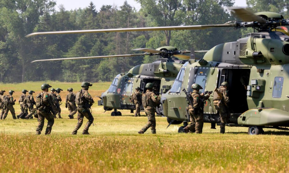 Soldiers in camouflage board a green military helicopter on a grassy airfield.