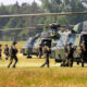 Soldiers in camouflage board a green military helicopter on a grassy airfield.