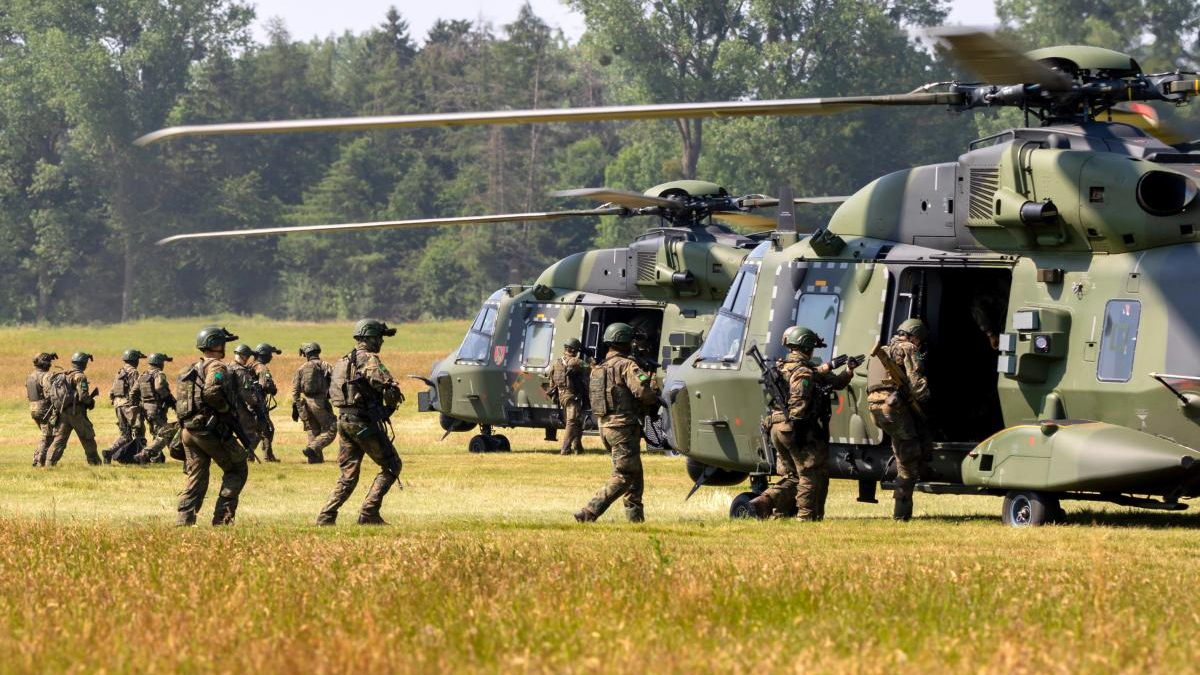 Soldiers in camouflage board a green military helicopter on a grassy airfield.