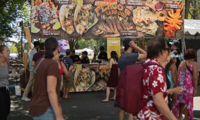 Busy outdoor food stall at a festival with a large banner featuring seafood tacos; people line up to order.