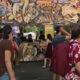 Busy outdoor food stall at a festival with a large banner featuring seafood tacos; people line up to order.