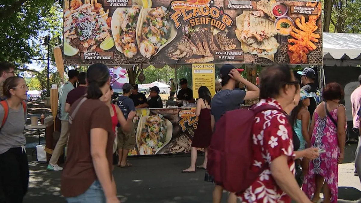 Busy outdoor food stall at a festival with a large banner featuring seafood tacos; people line up to order.