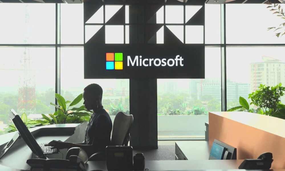 Silhouette of a person working at a curved reception desk in front of a large Microsoft sign with a windowed city view behind.