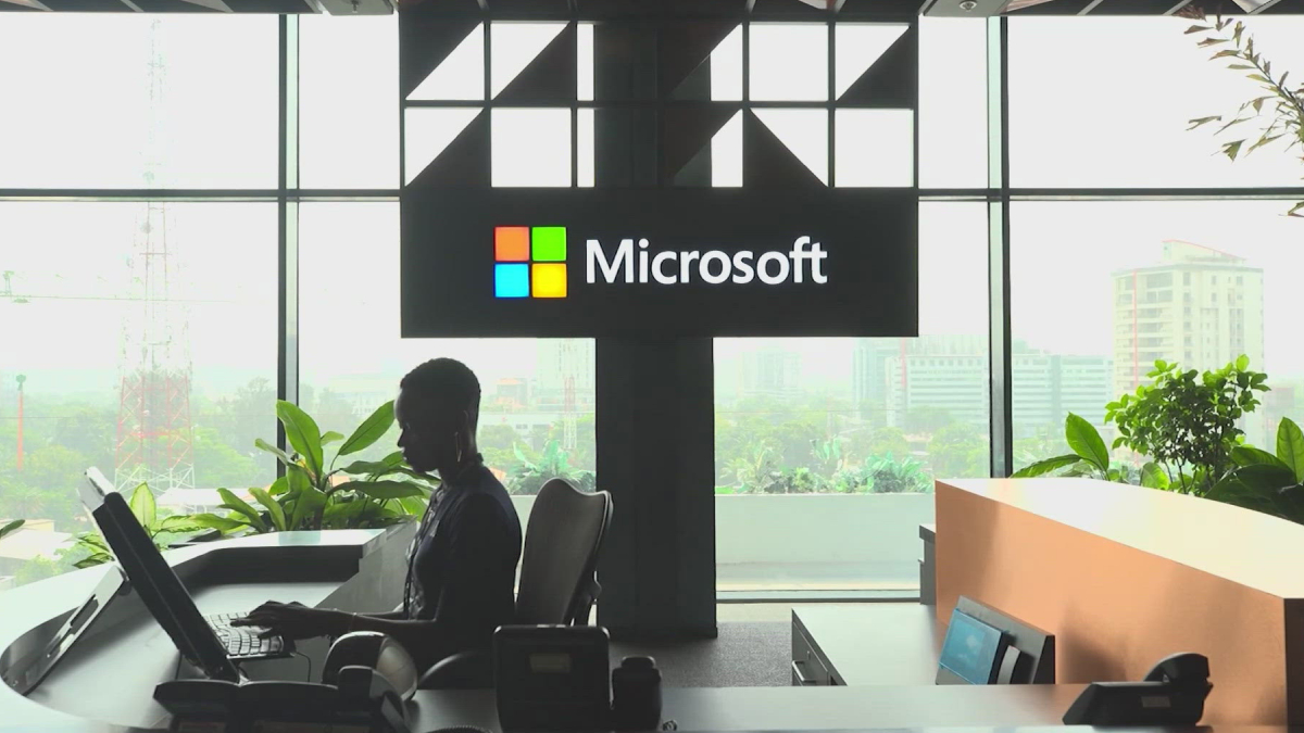 Silhouette of a person working at a curved reception desk in front of a large Microsoft sign with a windowed city view behind.