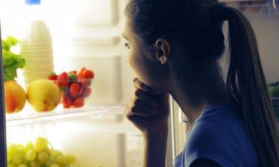 A woman stands in front of an open fridge, resting her chin on her hand and thinking about food choices.