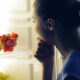 A woman stands in front of an open fridge, resting her chin on her hand and thinking about food choices.